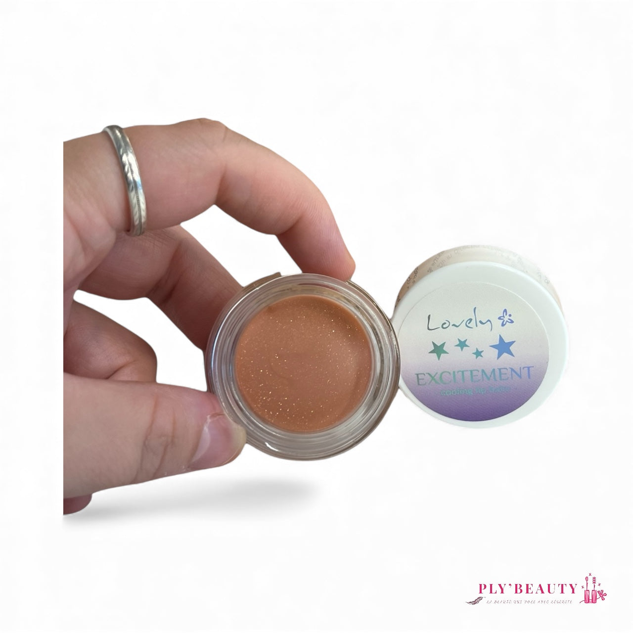 Baume à lèvres Lovely « Excitement » tenu entre les doigts, couleur nude pailletée dans un pot transparent, avec son couvercle blanc affichant des étoiles vertes et bleues. Logo Ply’Beauty en bas de l’image.