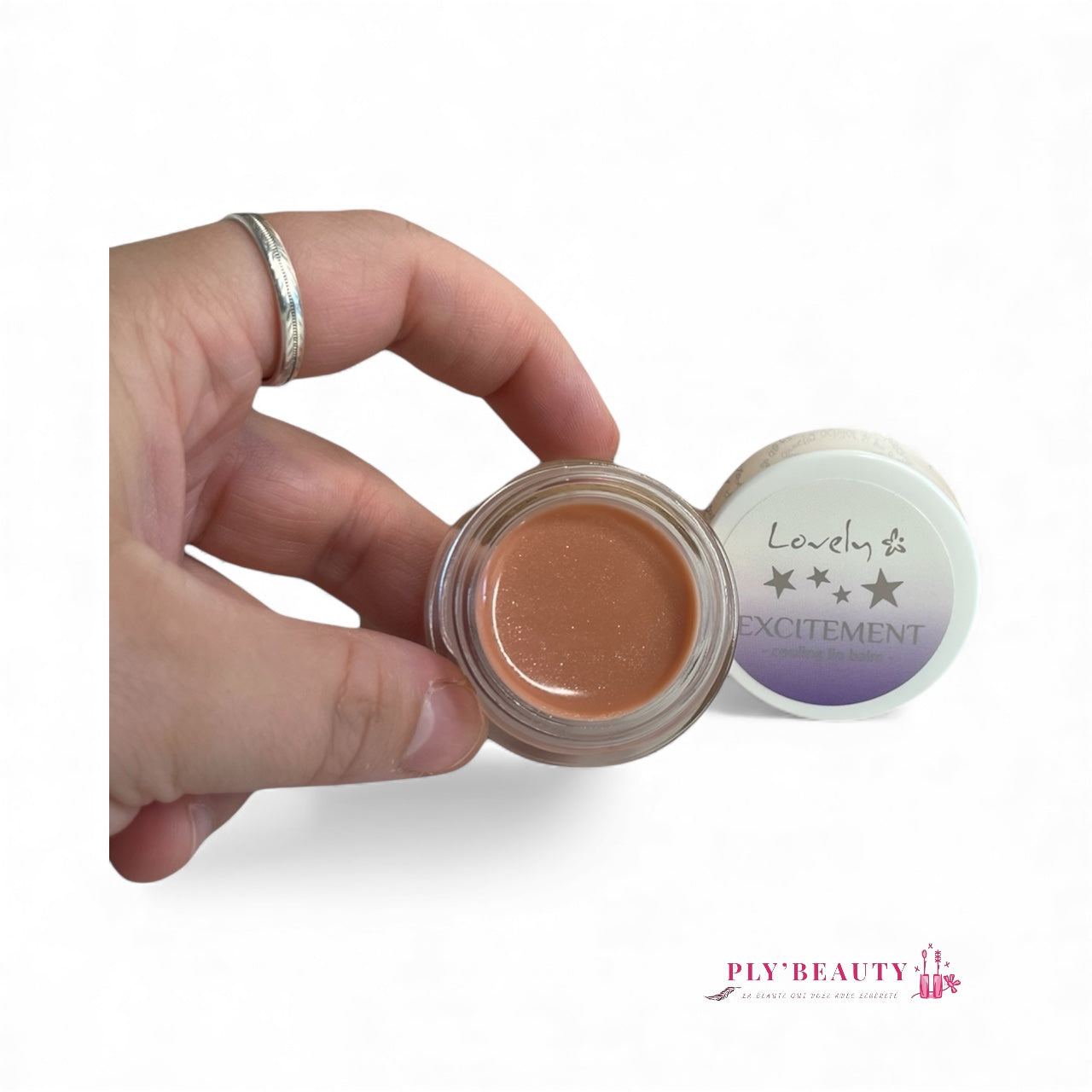 Baume à lèvres Lovely « Excitement » présenté ouvert dans la main, texture nude brillante dans un petit pot transparent, avec le couvercle blanc décoré d’étoiles violettes. Logo Ply’Beauty en bas.
