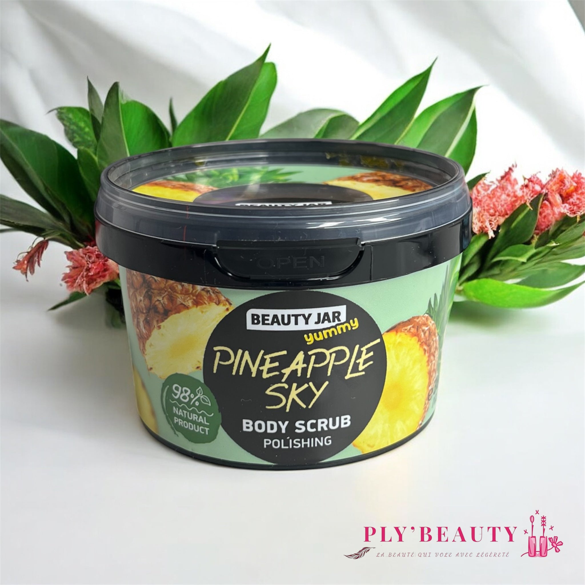 Gommage corps Beauty Jar Pineapple Sky 360 g, pot illustré d’ananas avec couvercle noir, 98% naturel, posé devant des feuilles vertes et fleurs tropicales.